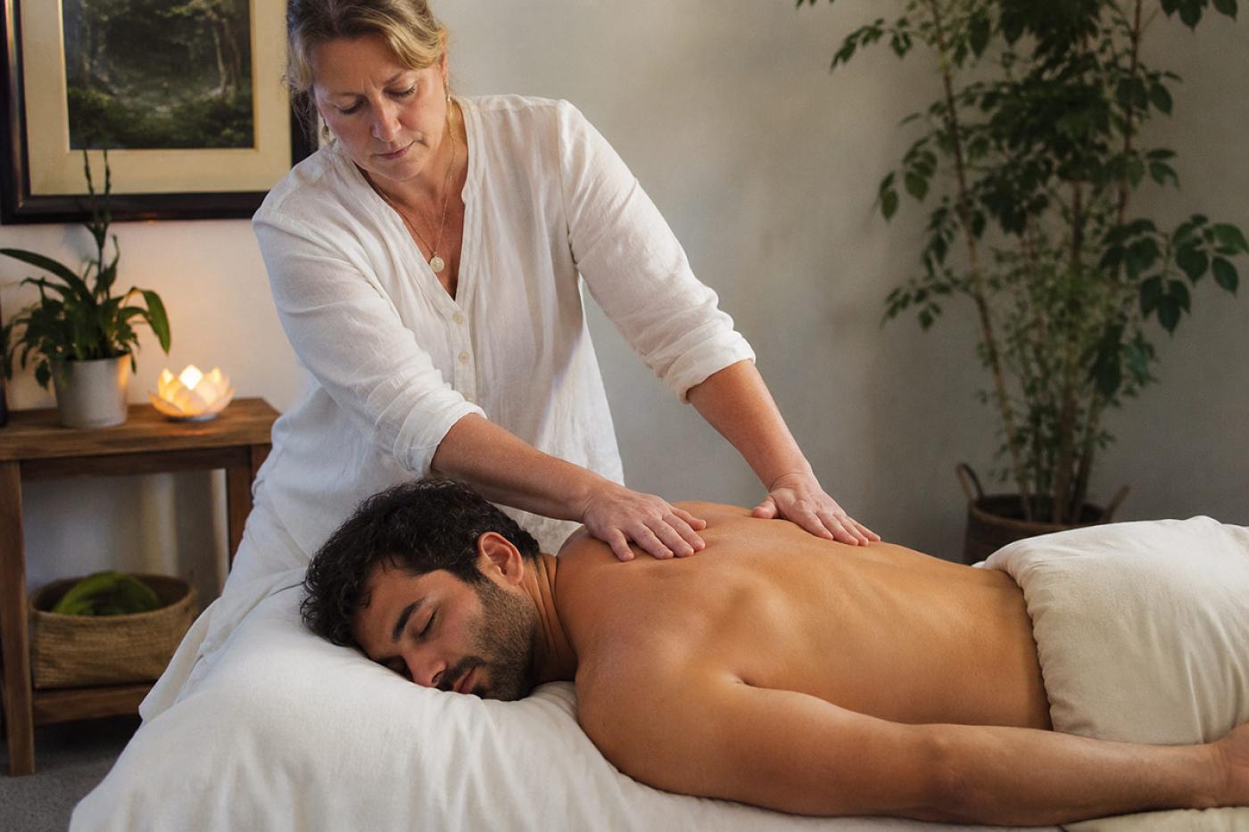 A therapist performs gentle massage bodywork on a relaxed client during a hybrid BCST and therapeutic massage session in a calm, softly lit treatment room.